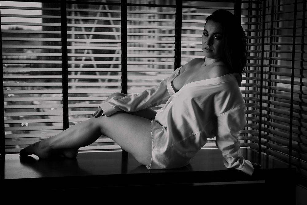 Artistic black and white boudoir photo of someone lounging in white clothing by a window with venetian blinds.