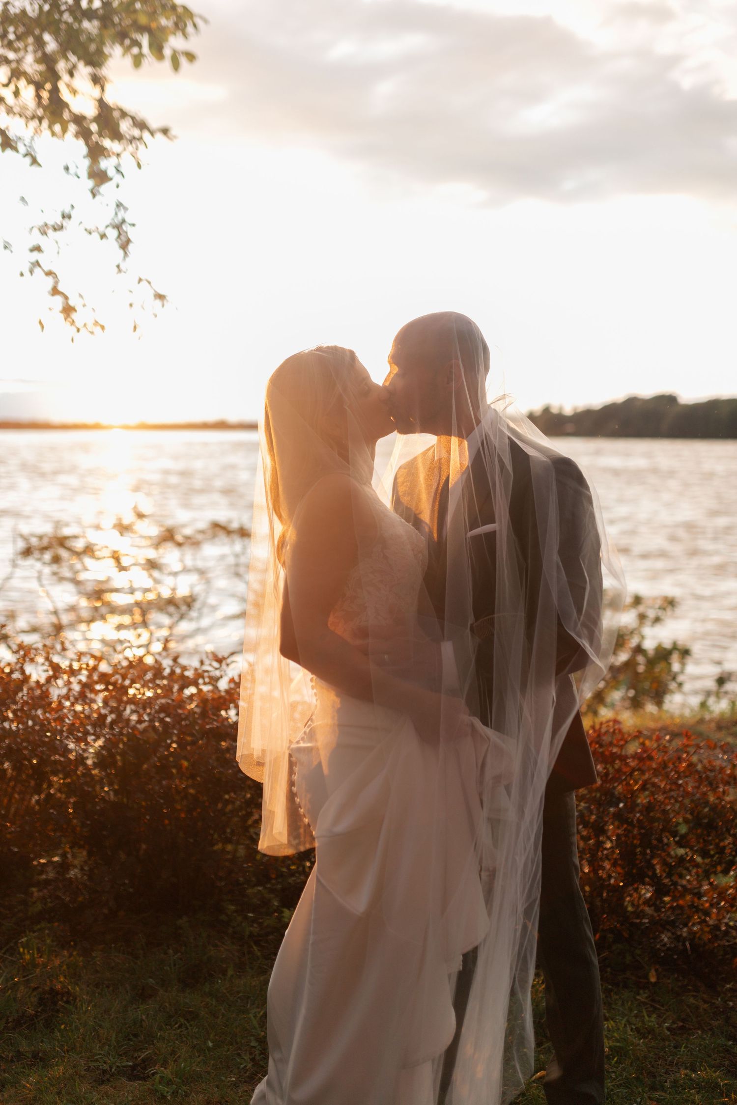 Bride and groom kissing under veil at golden sunset by a shimmering lake on their wedding day.