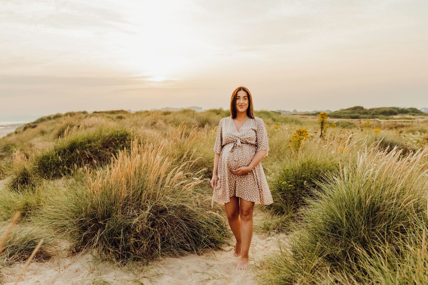 A pregnant woman walking along sandy Bournemouth beach, surrounded by tall beach grass.
