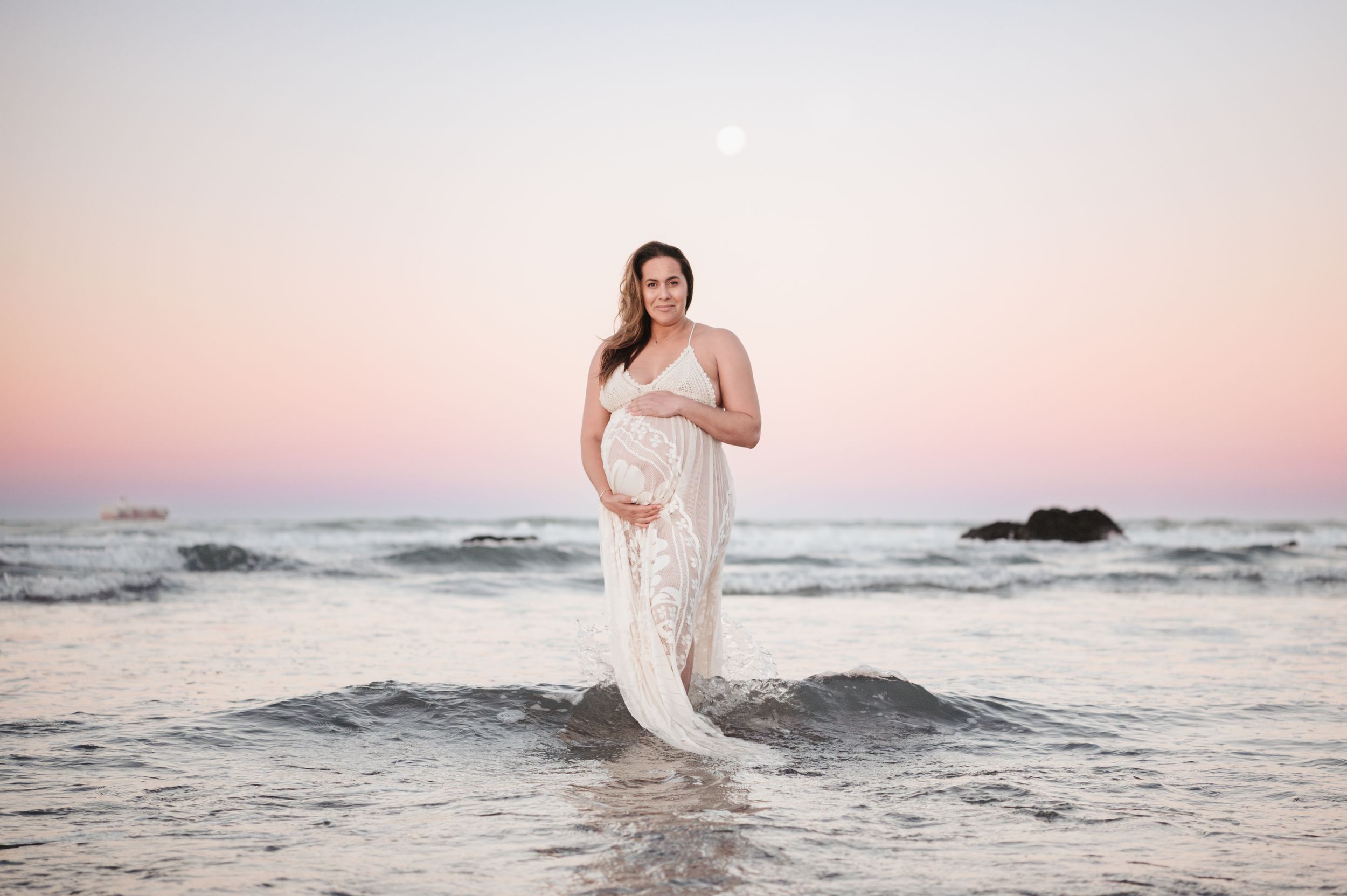 A person in a lace maternity gown stands at the edge of the ocean during a pink sunset.