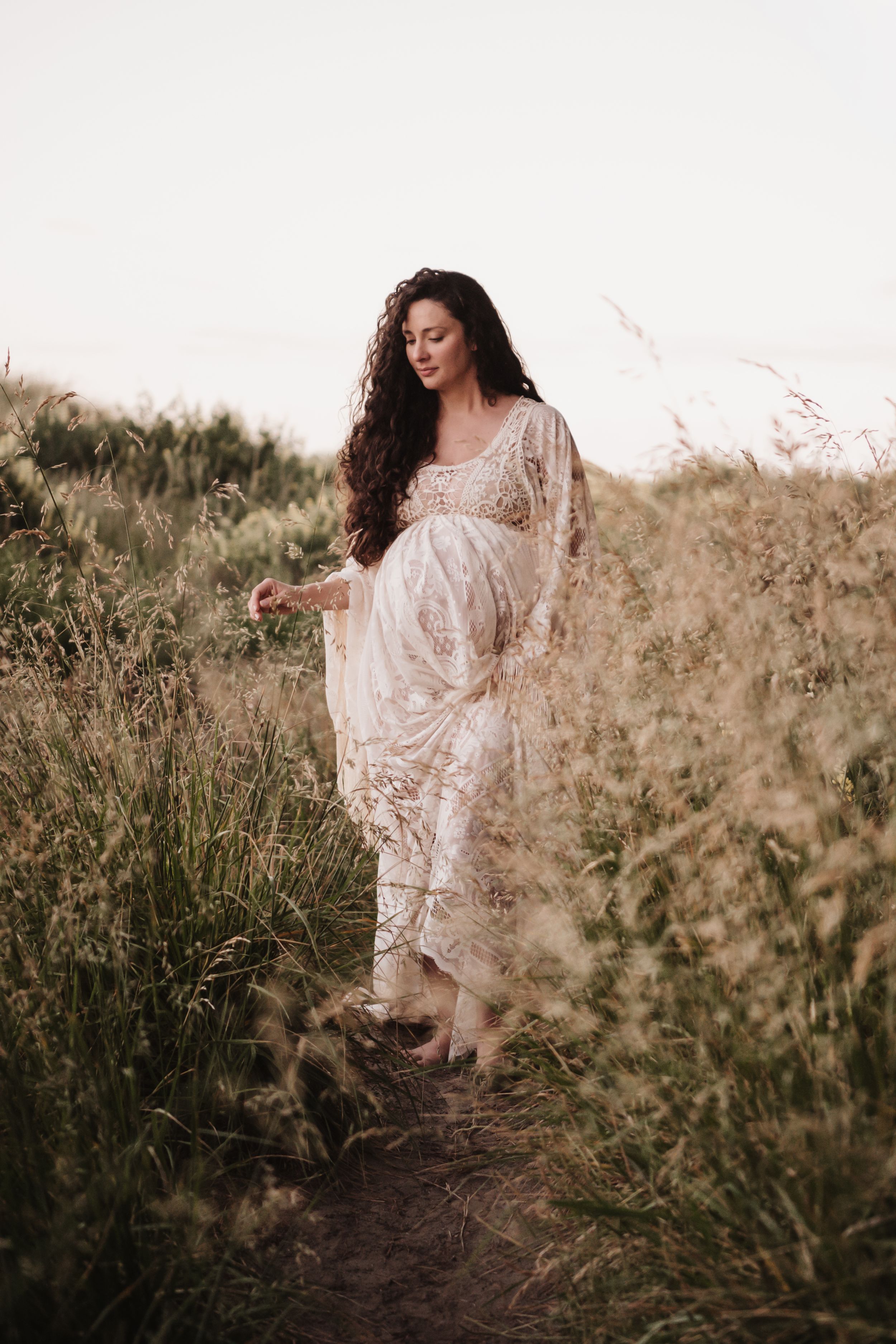 Expectant mother in lace dress stands among tall grass at golden hour.
