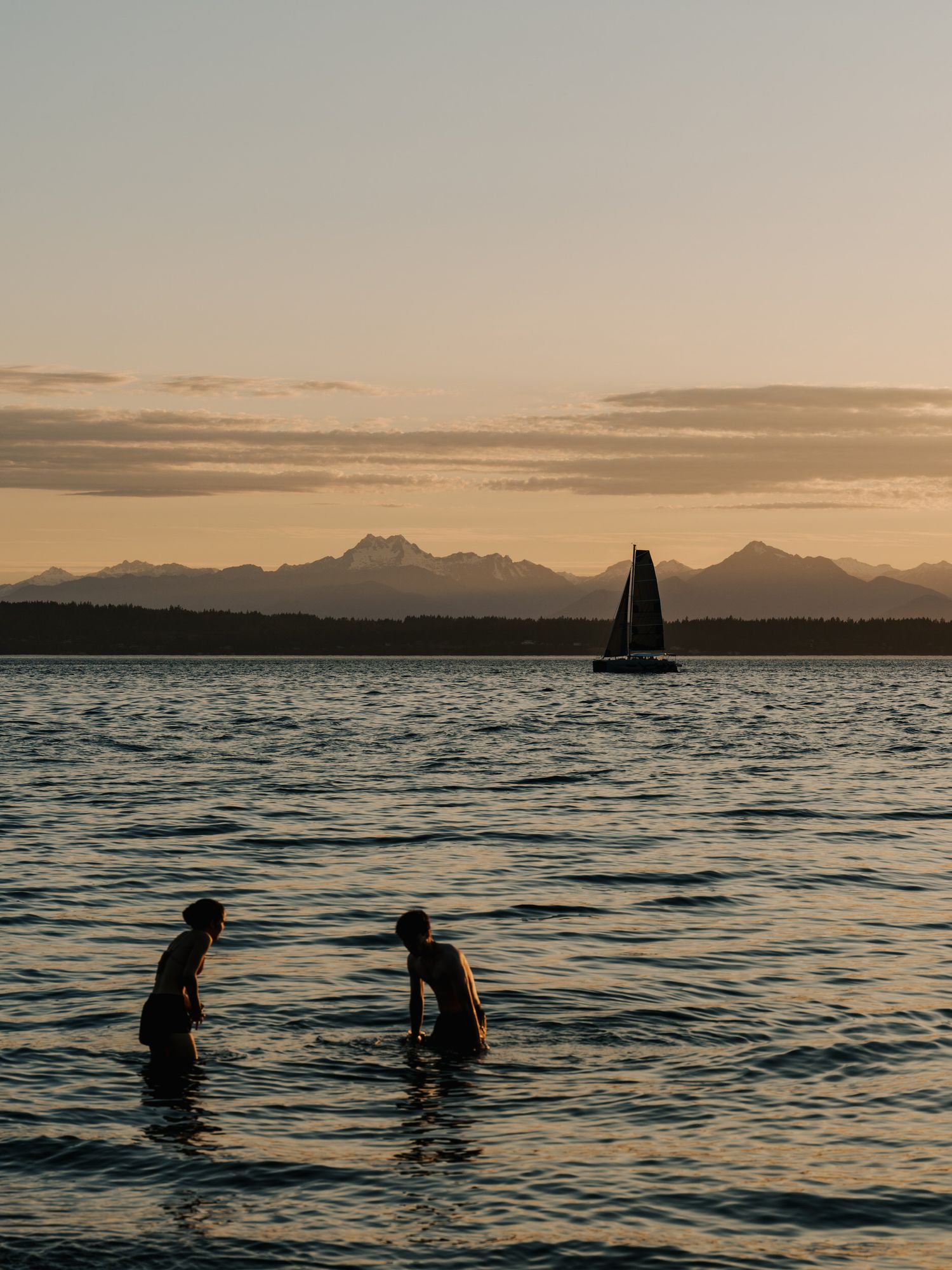 Silhouettes of swimmers in a lake at sunset with a sailboat and mountain range in the background.