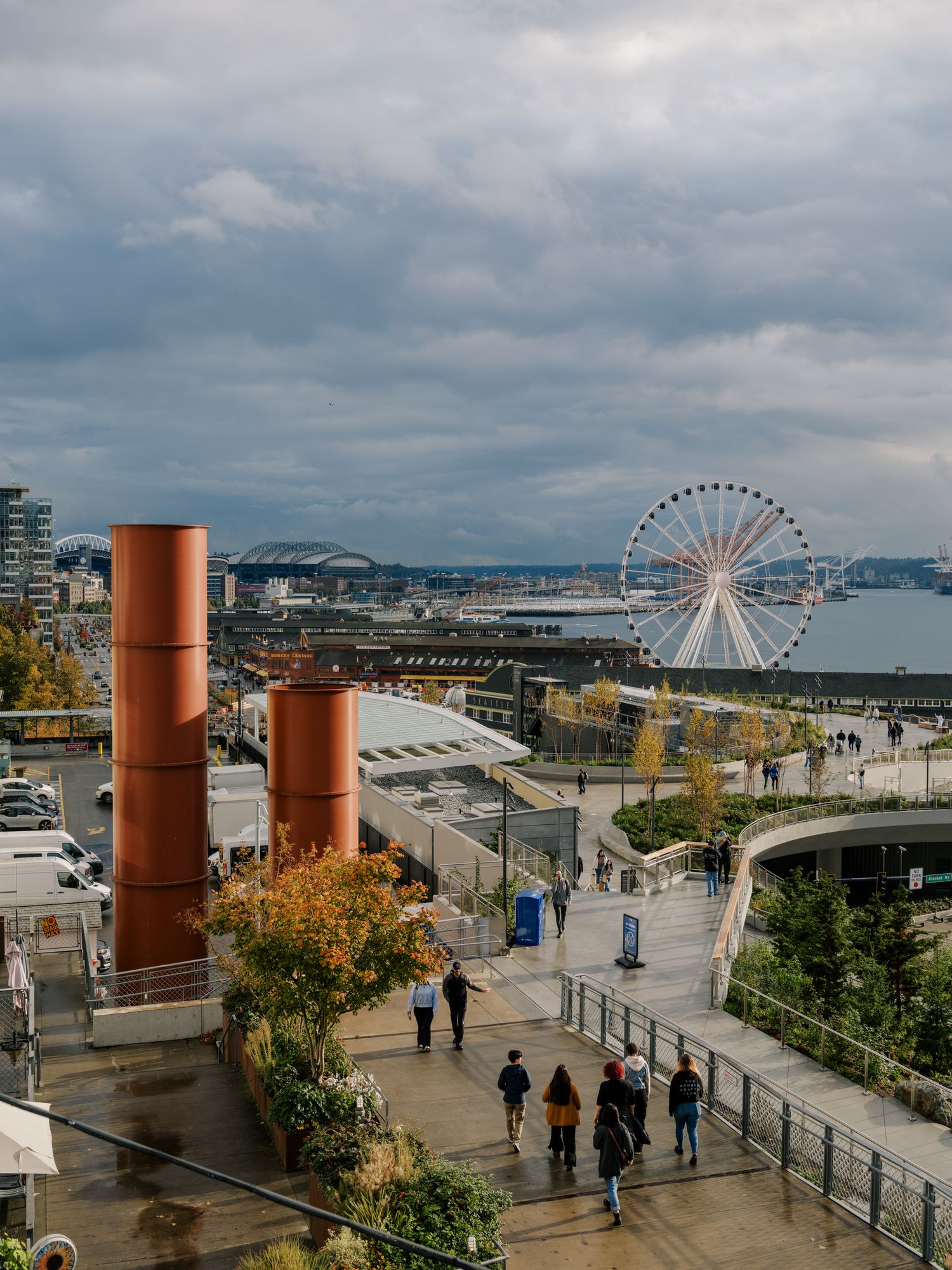 People walk along Seattle's waterfront with the Great Wheel and iconic orange smokestacks visible against cloudy skies.