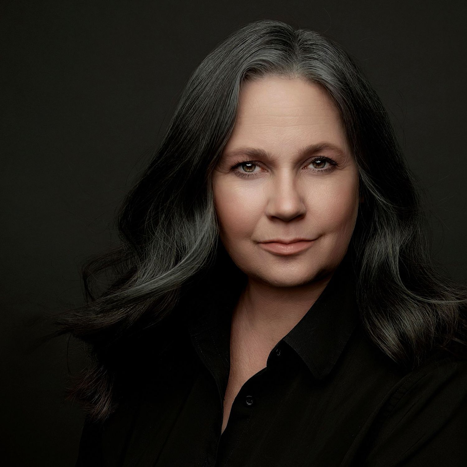 Professional studio portrait with dark background featuring soft lighting on a figure with flowing dark hair in black attire.