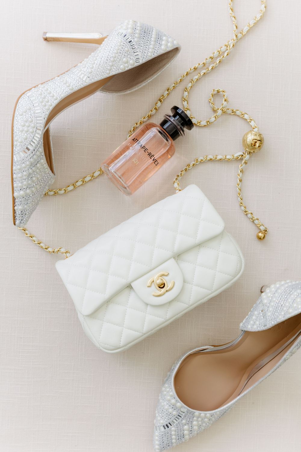 White quilted designer handbag with gold chain strap, sparkly heels, and pink perfume bottle on beige background.