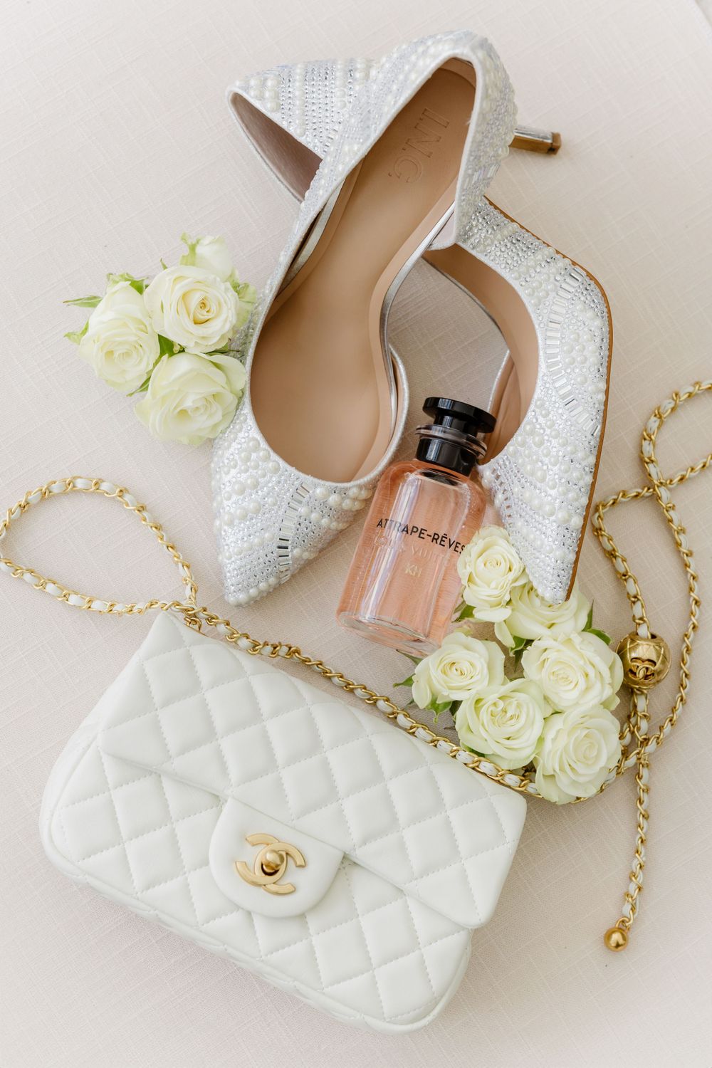Elegant white bridal accessories including sparkly heels, quilted purse, and white roses arranged on light background.