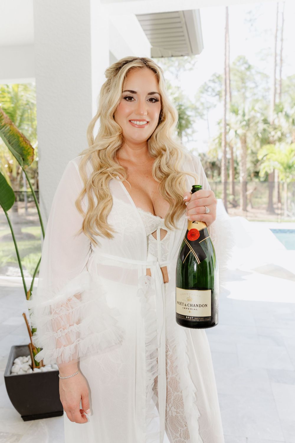 Celebratory moment with champagne bottle in white lace robe by tropical pool.