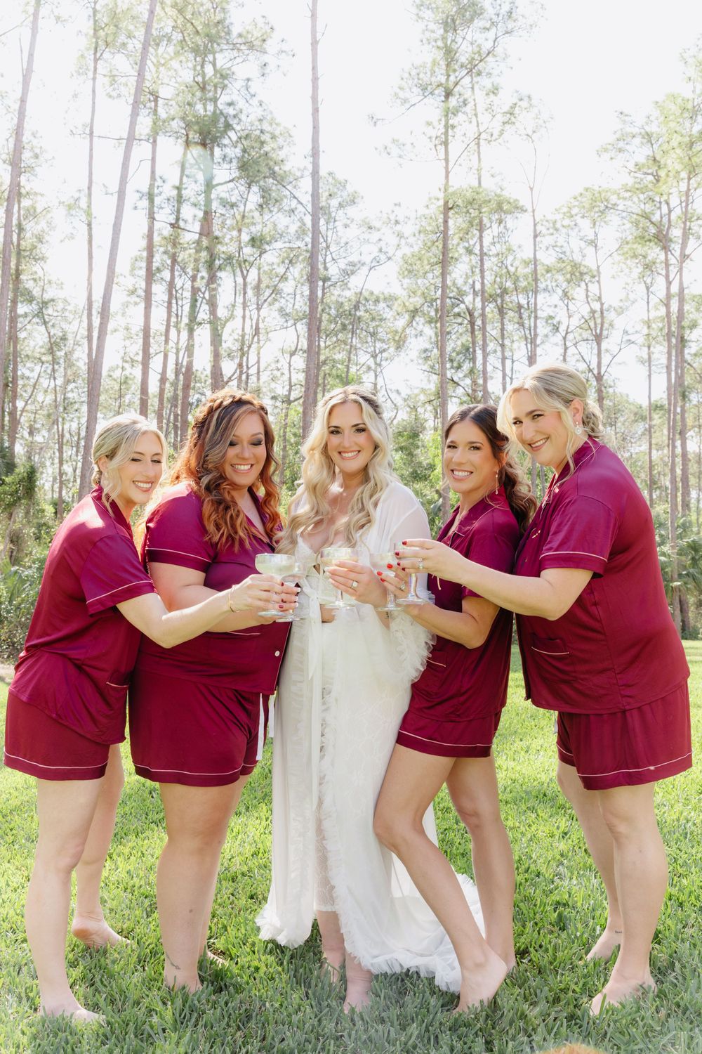 Bridal party in burgundy matching robes toasting outdoors among tall pine trees.