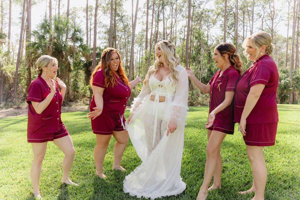 Wedding party in burgundy outfits celebrating with bride in white lace ensemble outdoors.