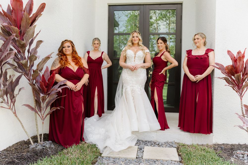 Wedding party poses outdoors with bridesmaids in burgundy gowns around bride in white dress.