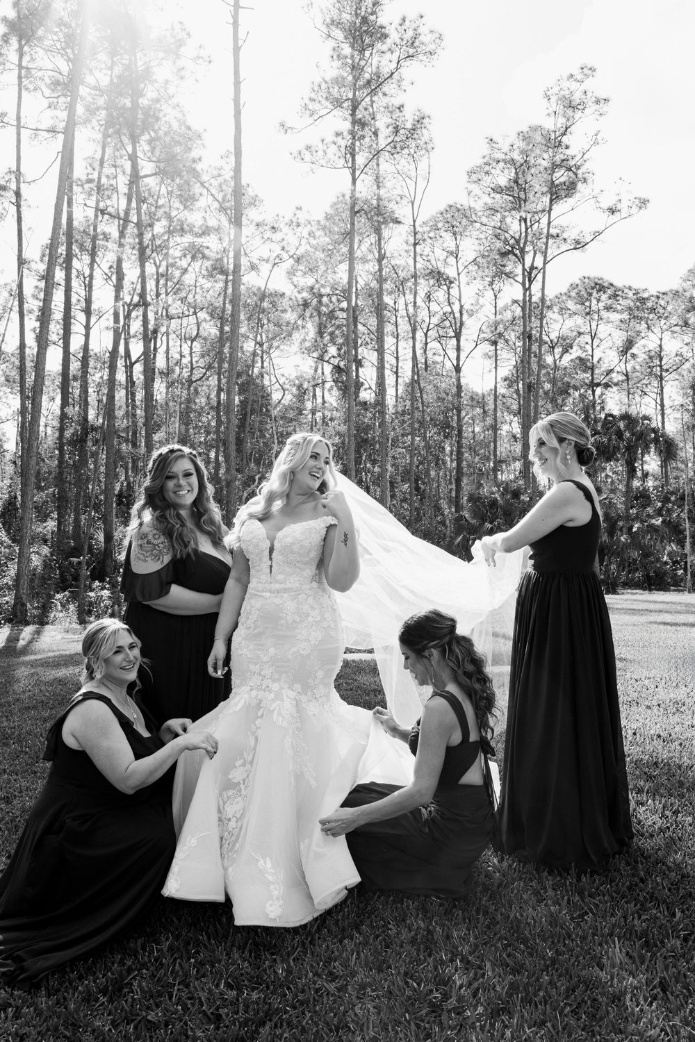 Black and white photo of bridesmaids helping with wedding dress outdoors.