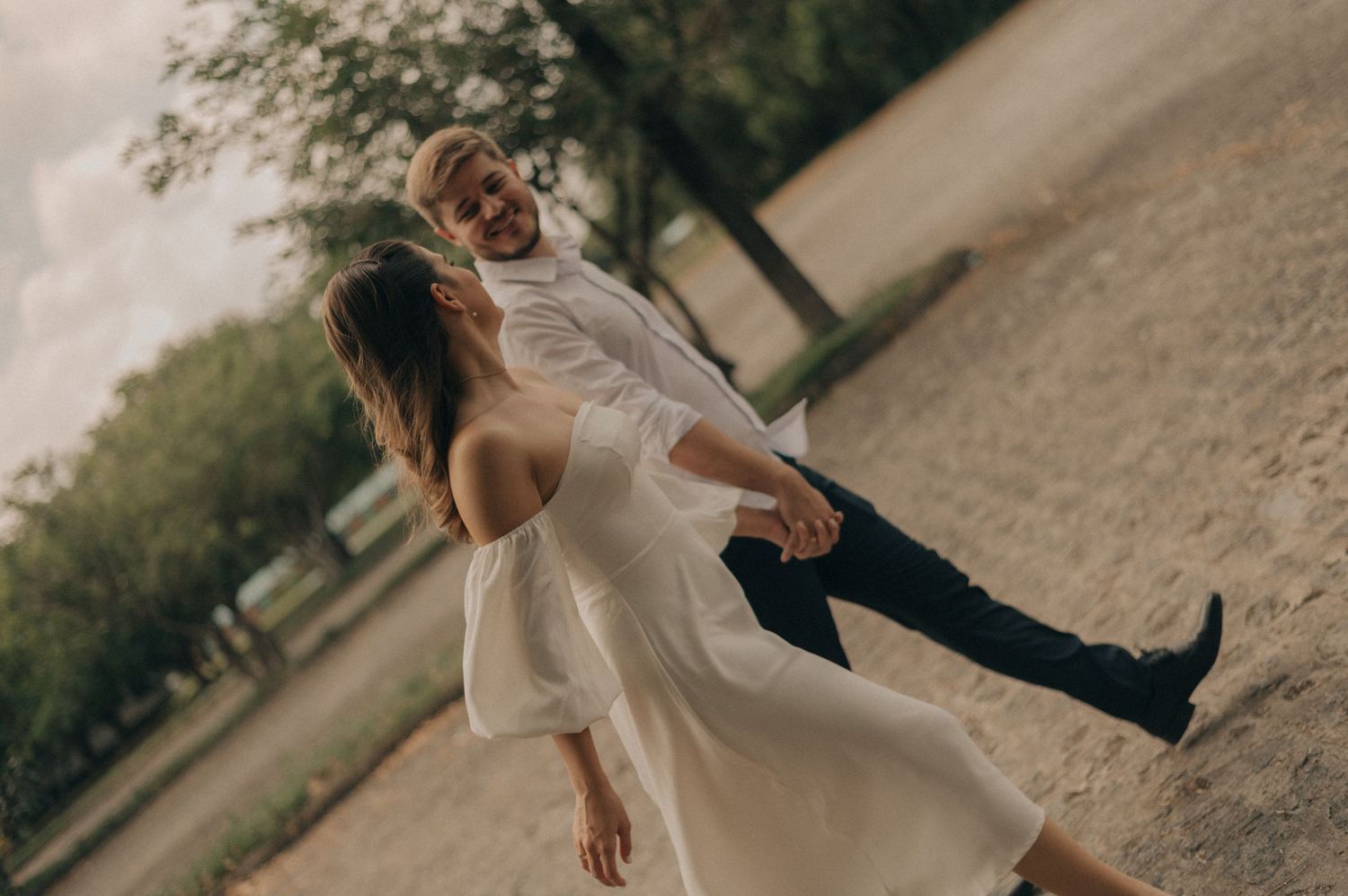 Playful couple in white clothing dancing outdoors on dirt path.