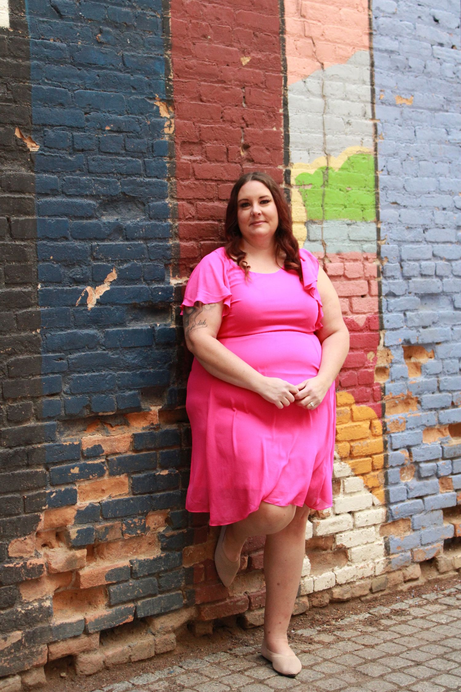 Someone wearing a pink flutter sleeve dress stands in front of a painted brick wall with multicolored sections.
