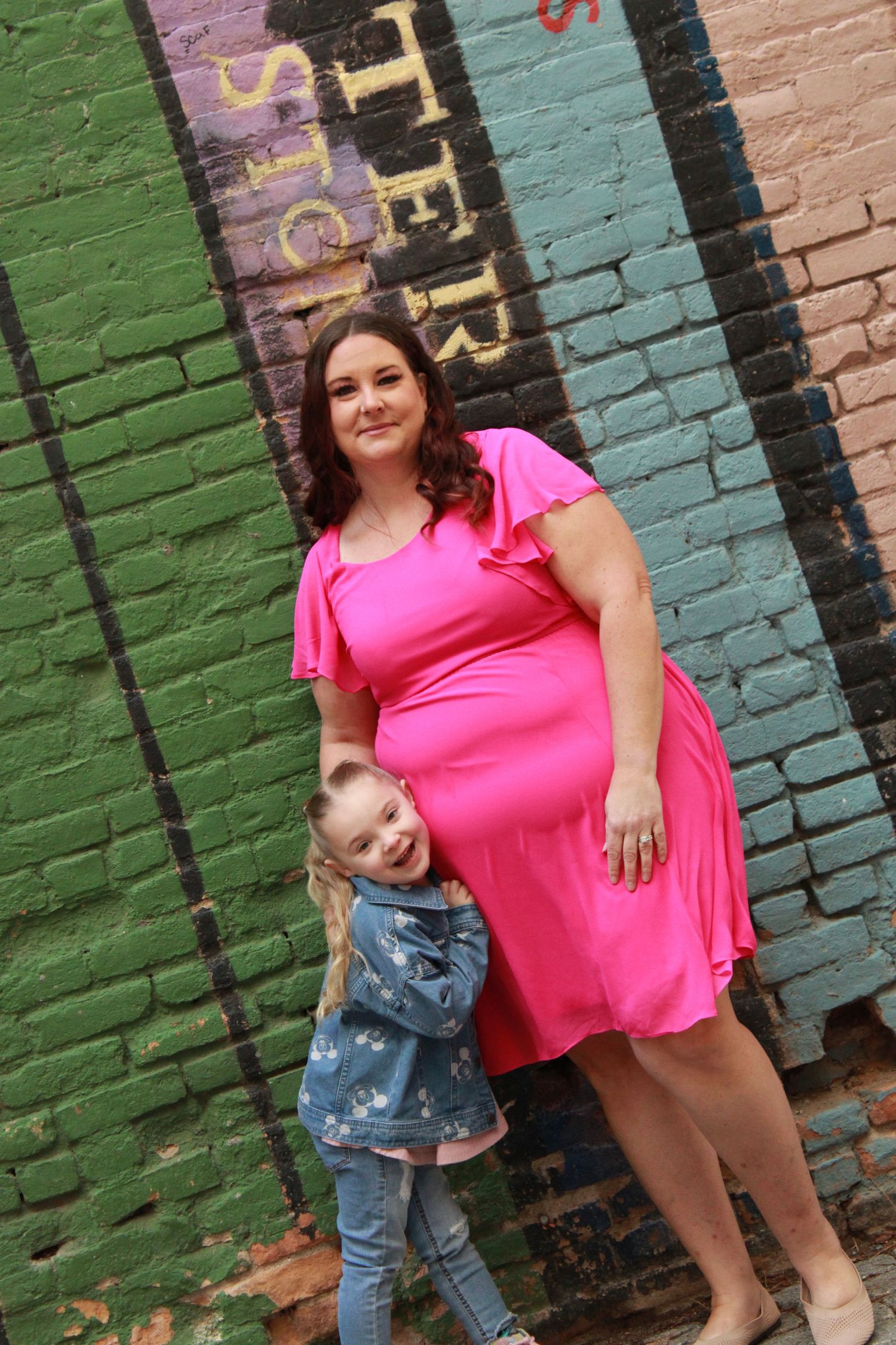 A bright pink dress stands out against multicolored brick wall as a small figure peeks out playfully.