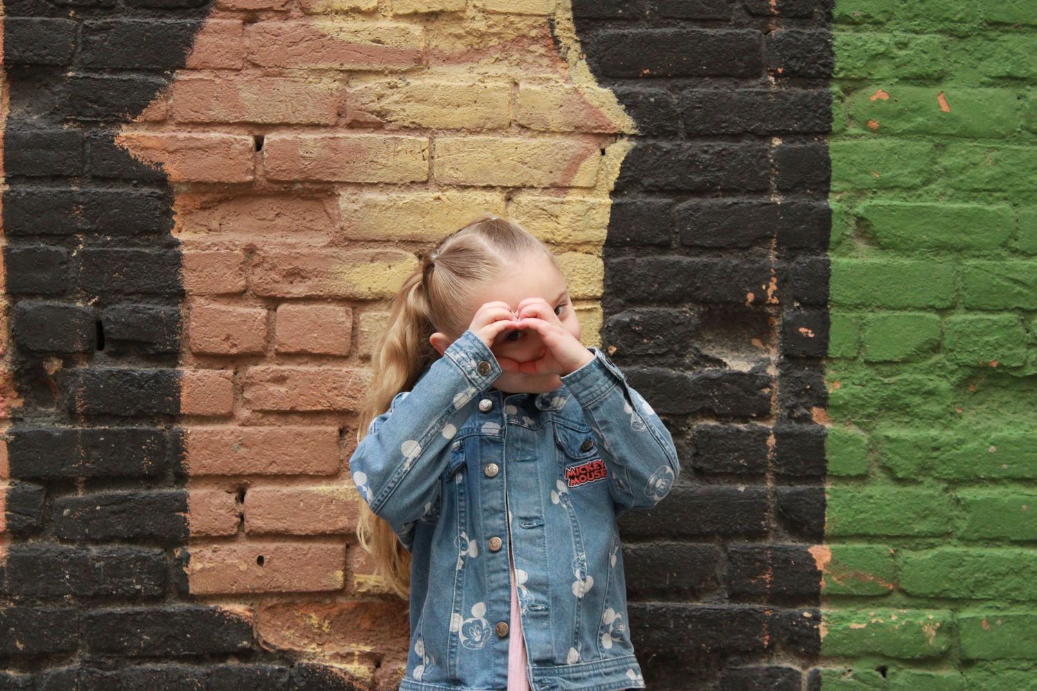 A small figure in denim jacket plays peek-a-boo against a colorful brick wall backdrop.