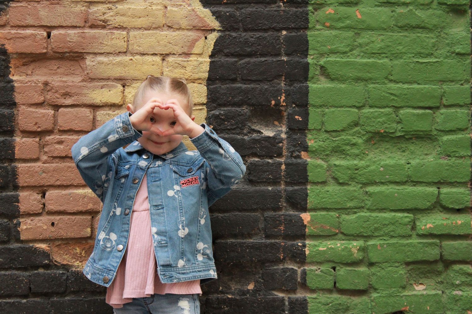 A playful pose against vibrant painted brick walls shows someone covering their eyes with both hands.