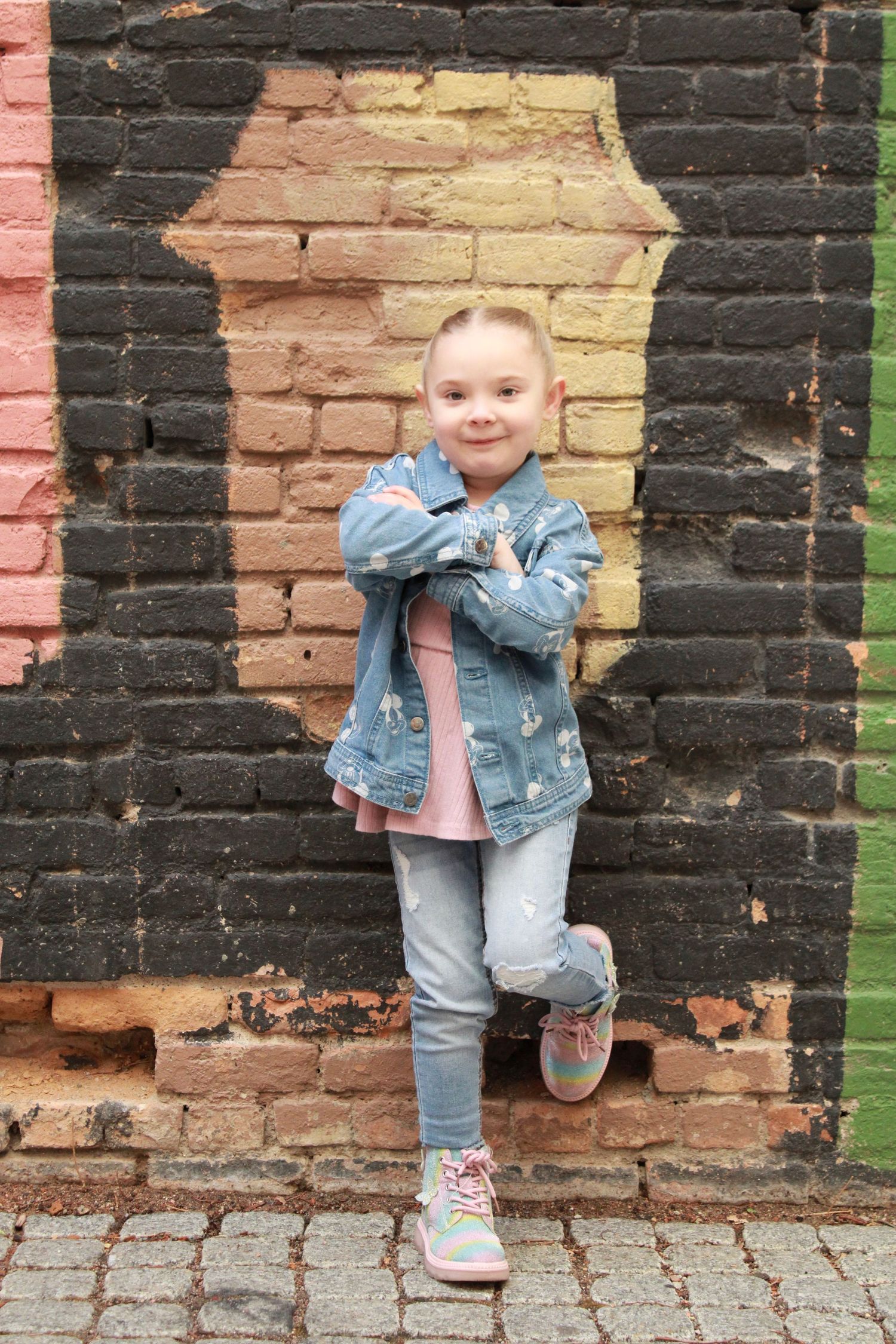 A small figure strikes a cute pose in denim jacket and jeans against colorful brick wall background.