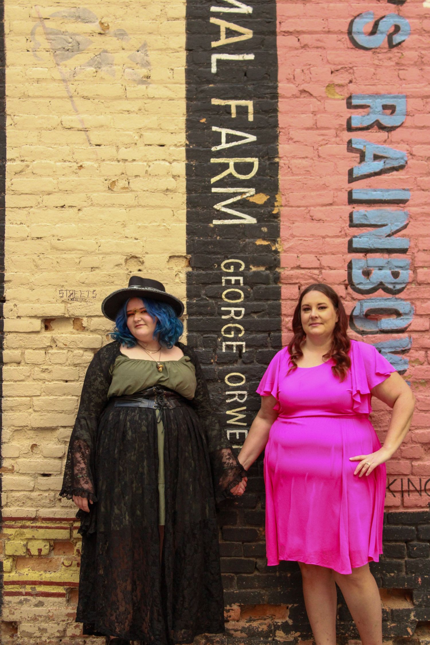Two people in contrasting outfits pose against a painted brick wall with text.