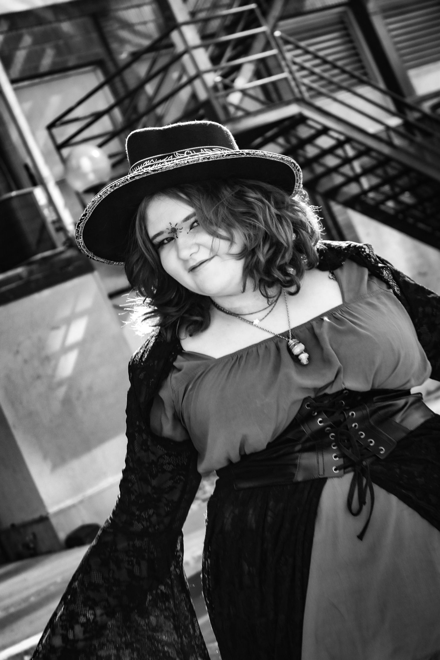 A black and white gothic portrait featuring a black hat and lace sleeves in an industrial setting.