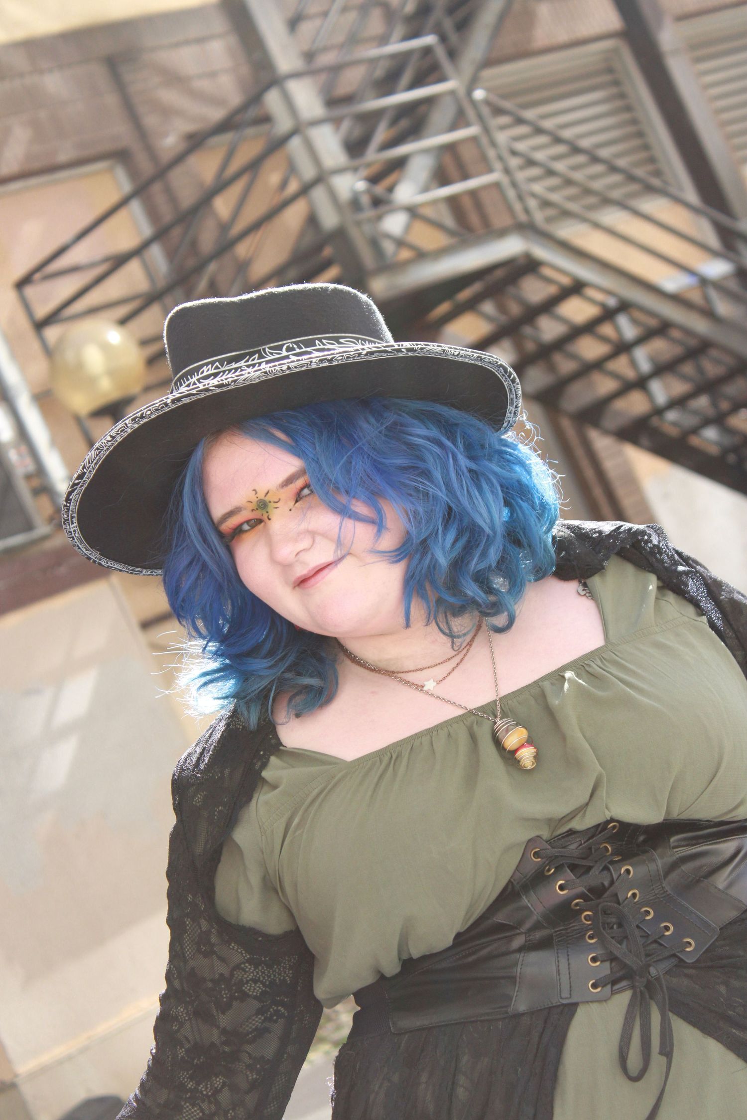 A gothic style photo showcasing a black hat and blue hair against an industrial backdrop with stairs.