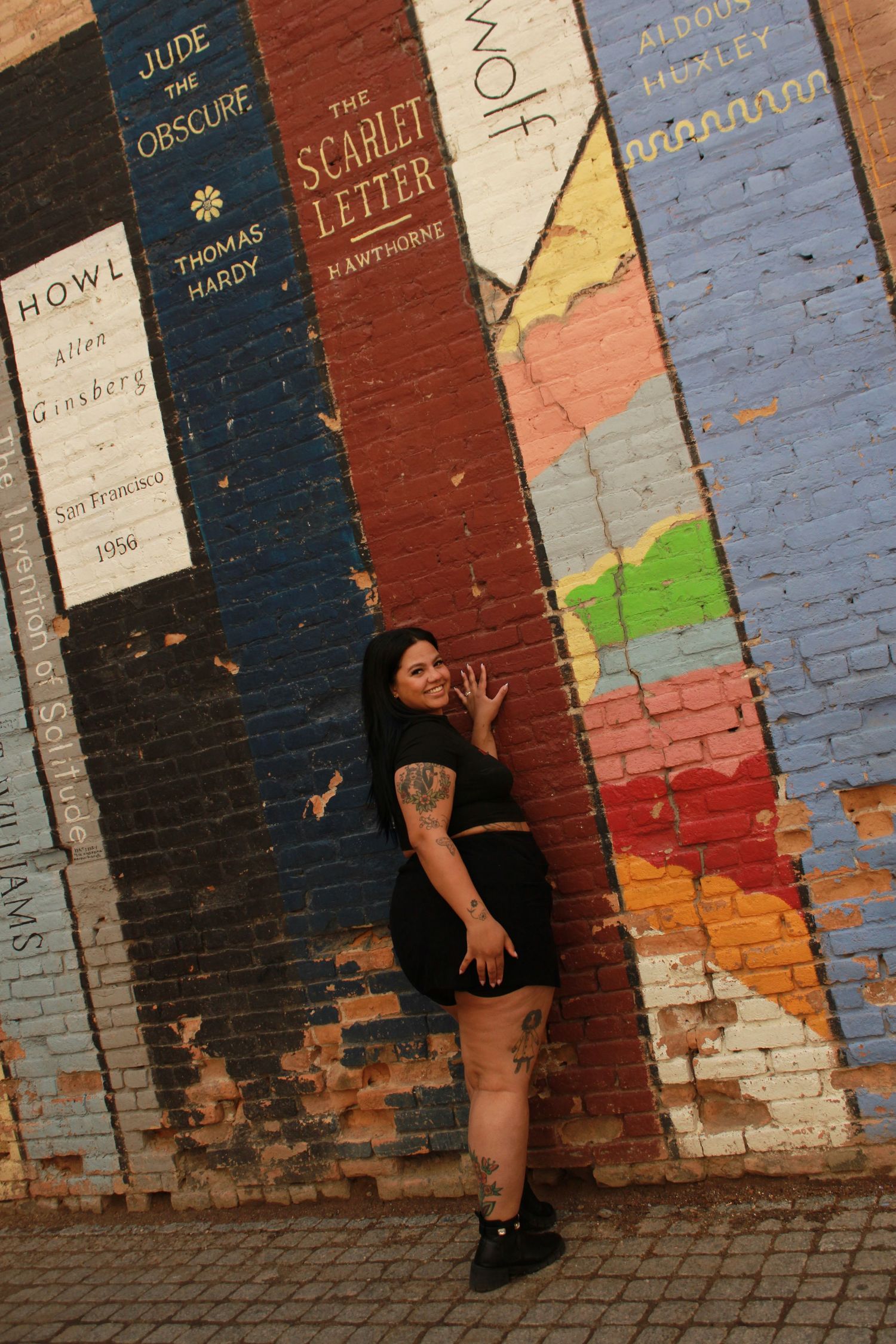 Artistic wall mural featuring classic book spines painted on brick with person posing against colorful design.