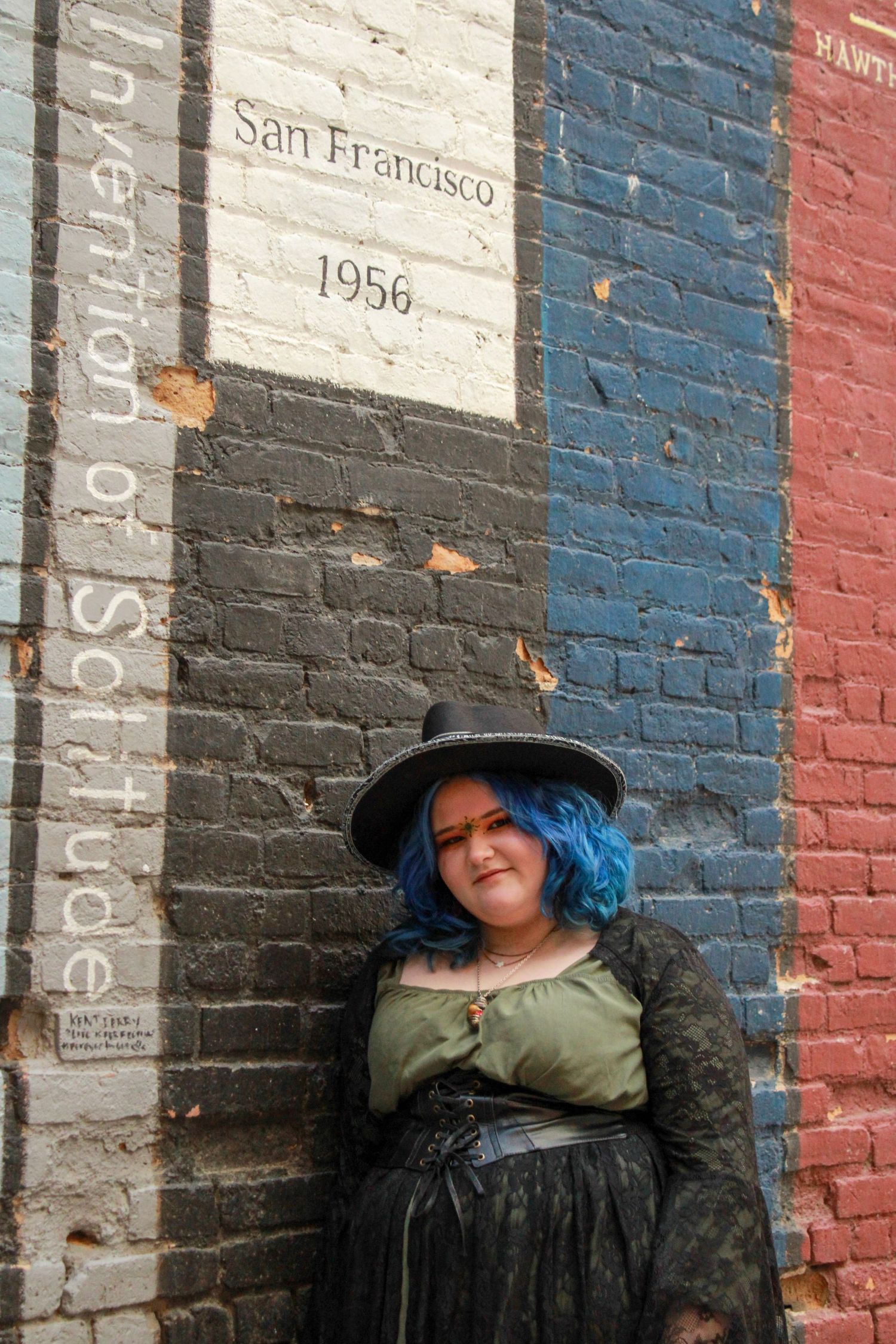 A figure in a black hat and green top poses against a colorful brick wall with San Francisco 1956 text visible.