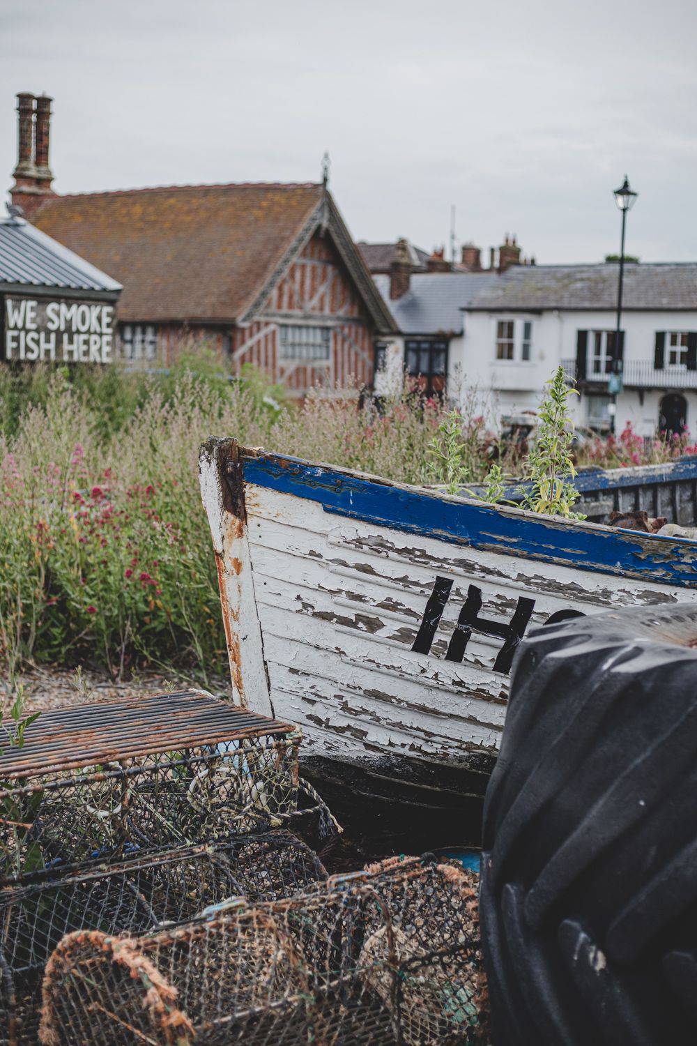 Weathered white and blue boat with rustic buildings and 'We Smoke Fish Here' sign in background. Aldeburgh Suffolk
