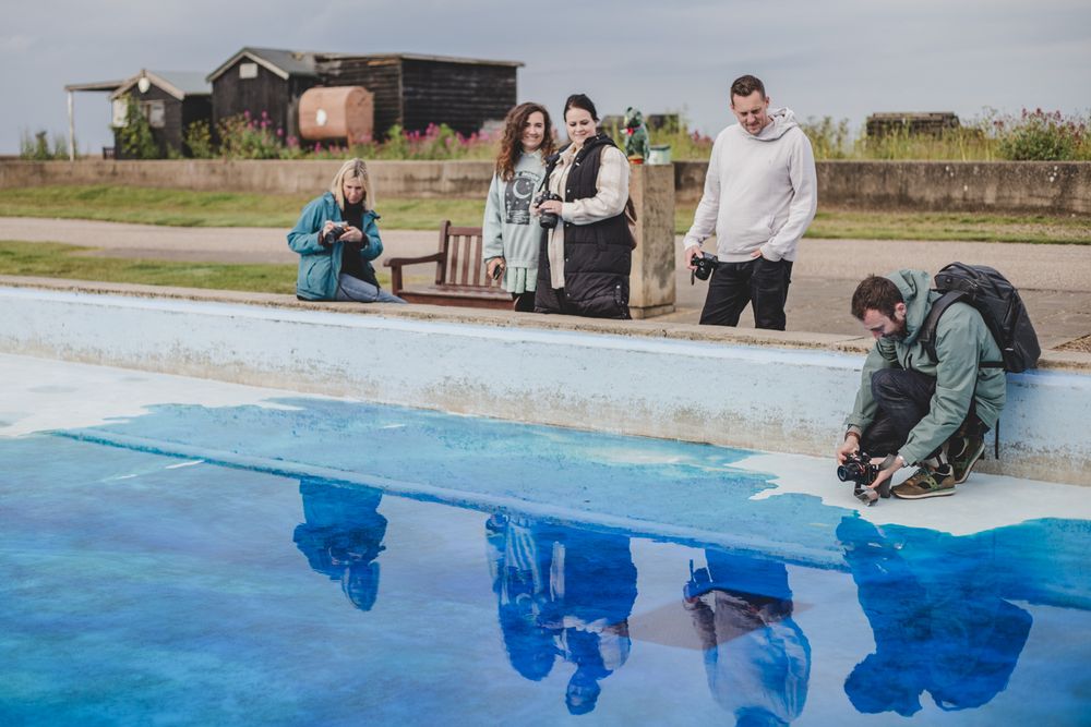 People gathered around a bright blue swimming pool on an overcast day.