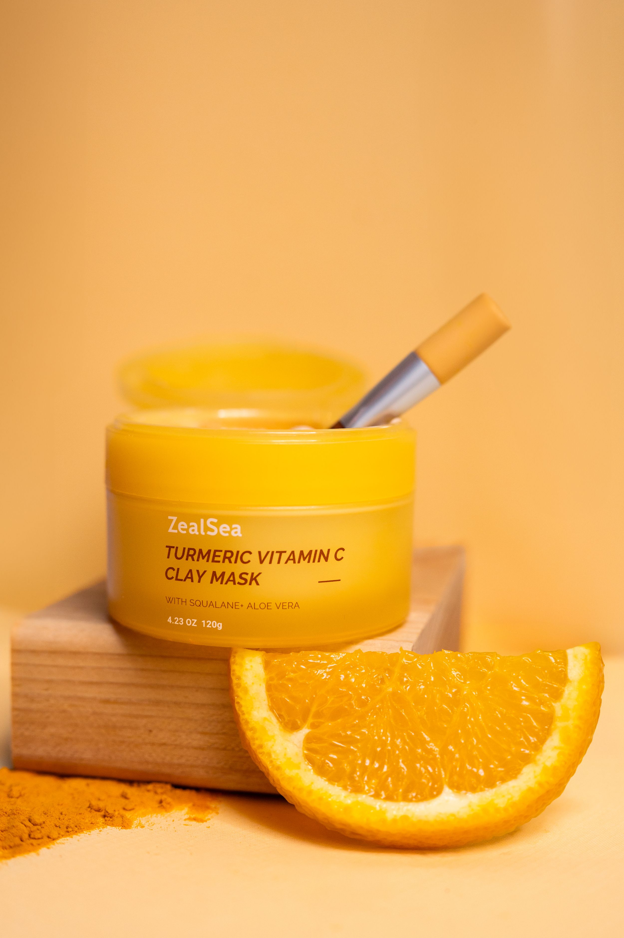 Yellow jar of turmeric vitamin C clay mask displayed with fresh orange slice and turmeric powder on wooden surface.