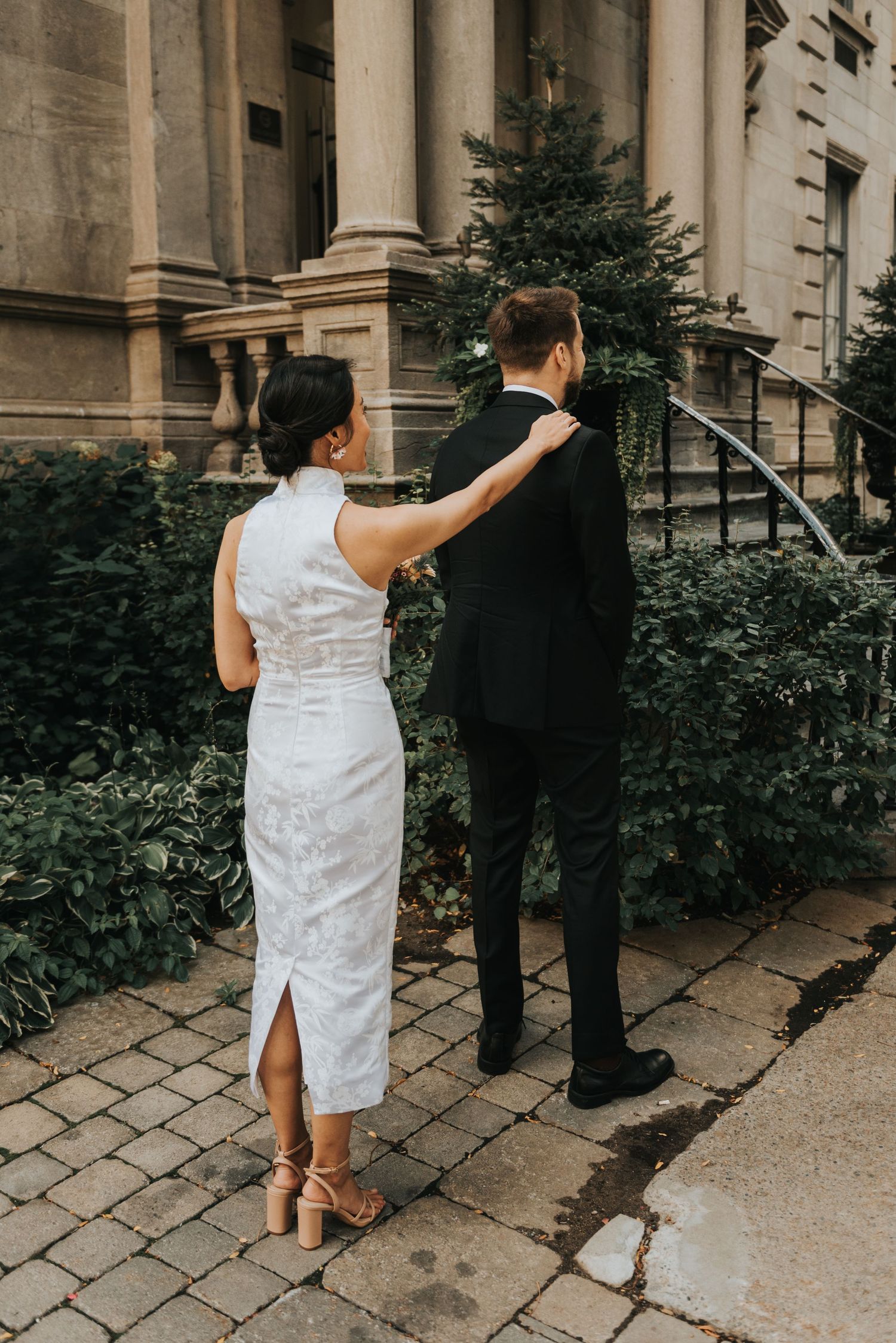 A couple in formal attire stands on a stone path outside a historic building with pillars and greenery.