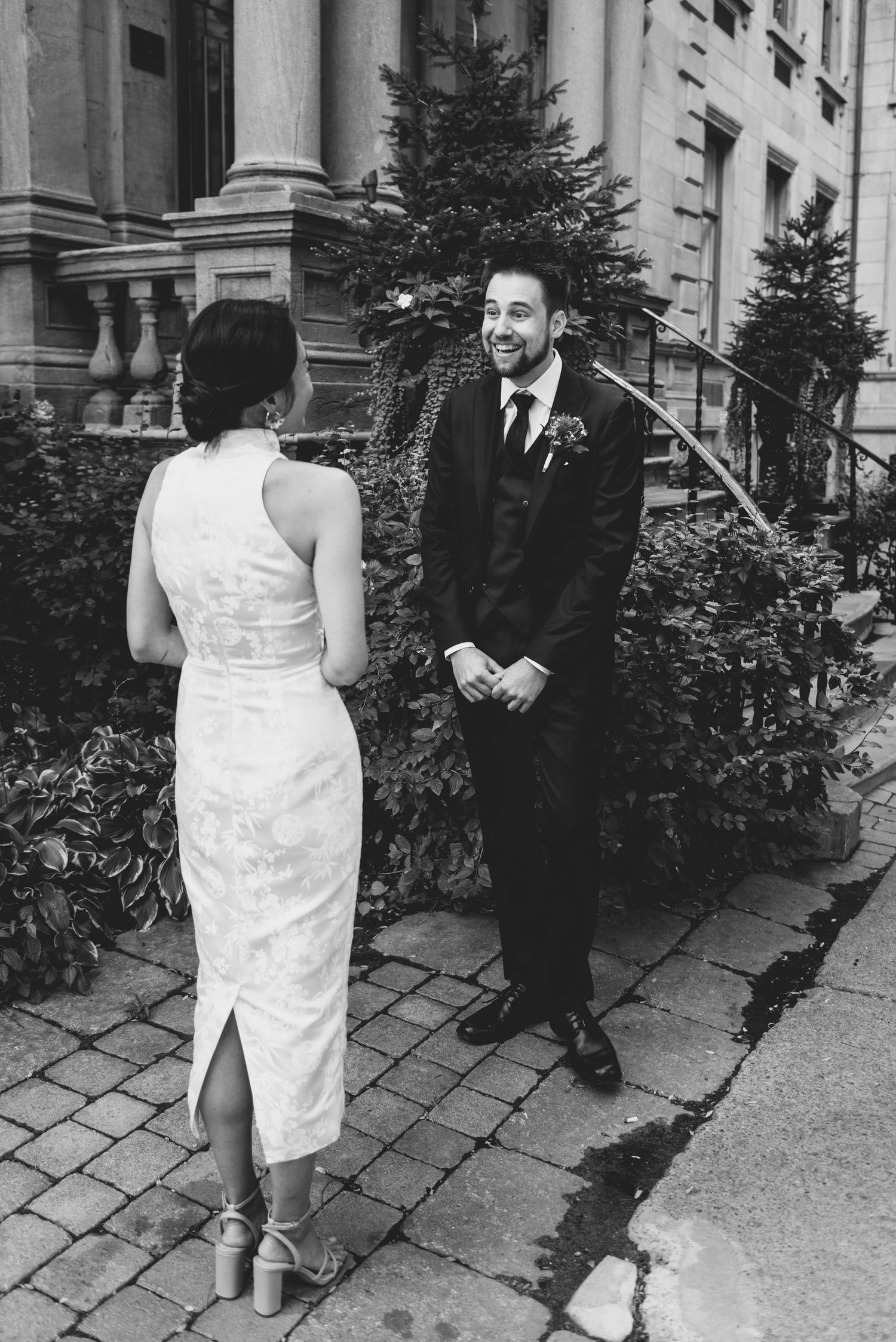 A couple shares their first look moment in black and white outside a historic building with evergreen trees.