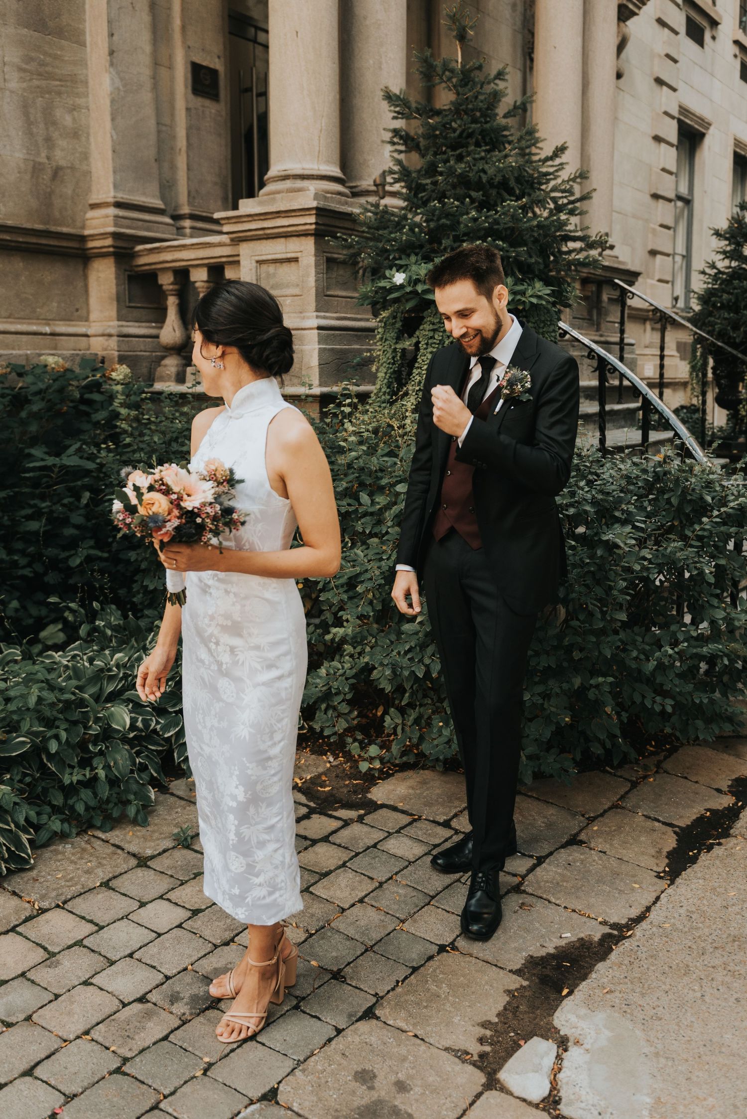 A couple in formal wedding attire stand on a stone path outside a historic building with pillars and greenery.