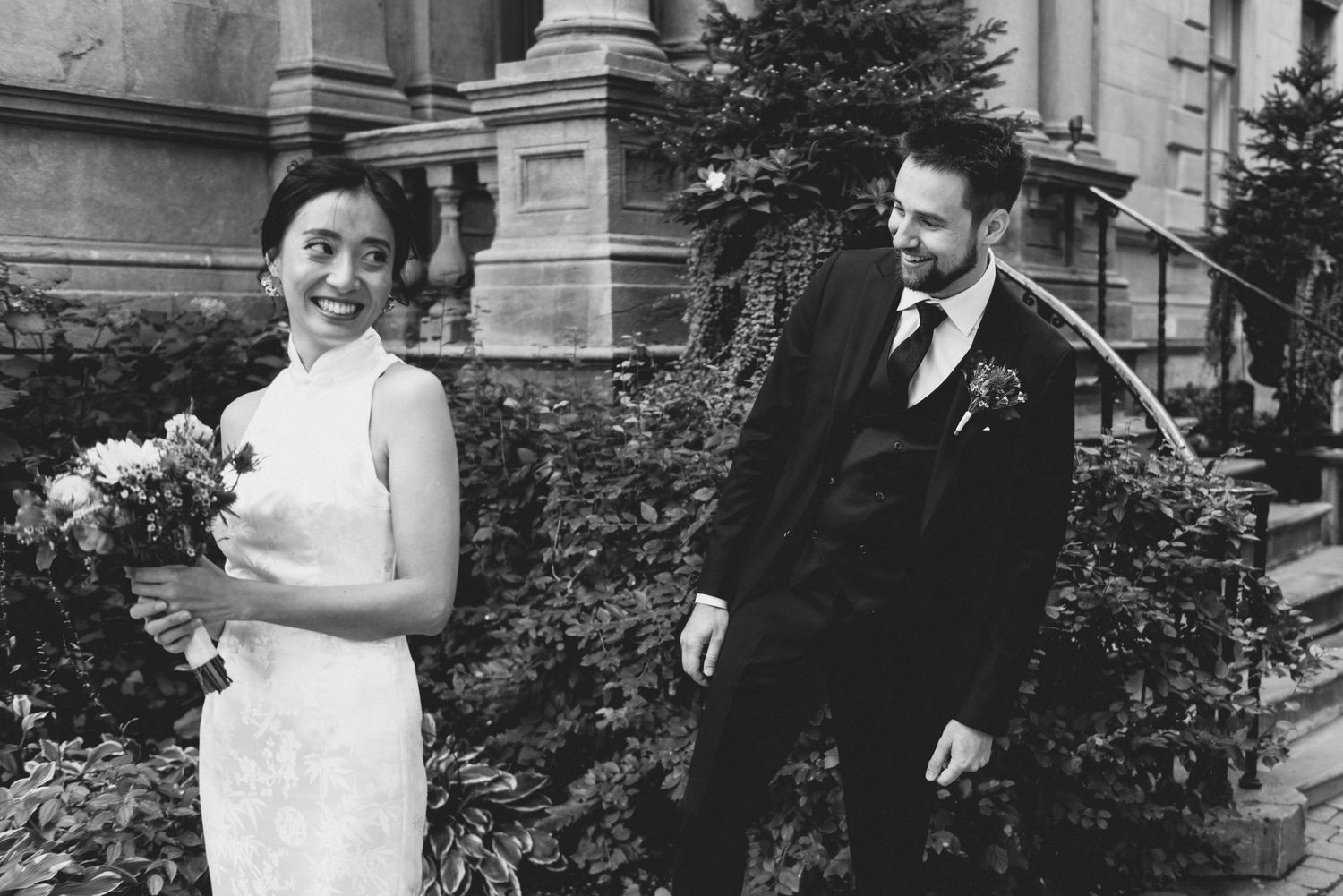 A black and white photograph of a bride and groom standing together in formal attire outside a stately building.