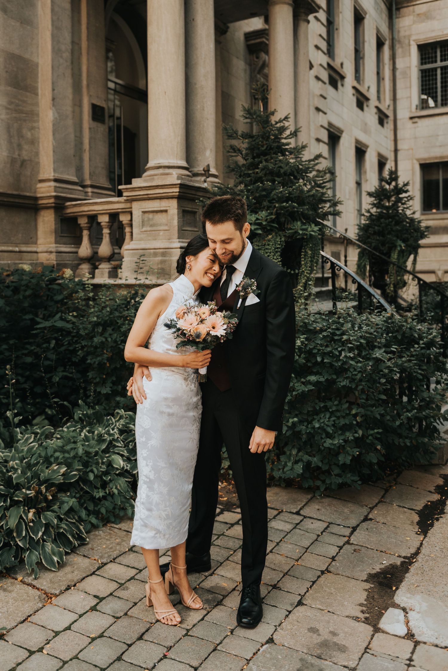 A romantic wedding couple shares an intimate moment outside a historic building with white columns and stone pathways.