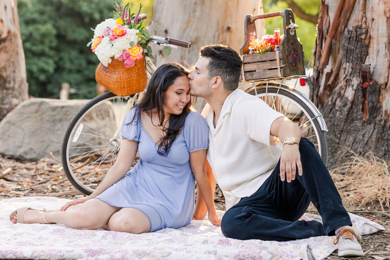 Couple sits on picnic blanket near vintage bicycle decorated with floral arrangements in pumpkin vases.