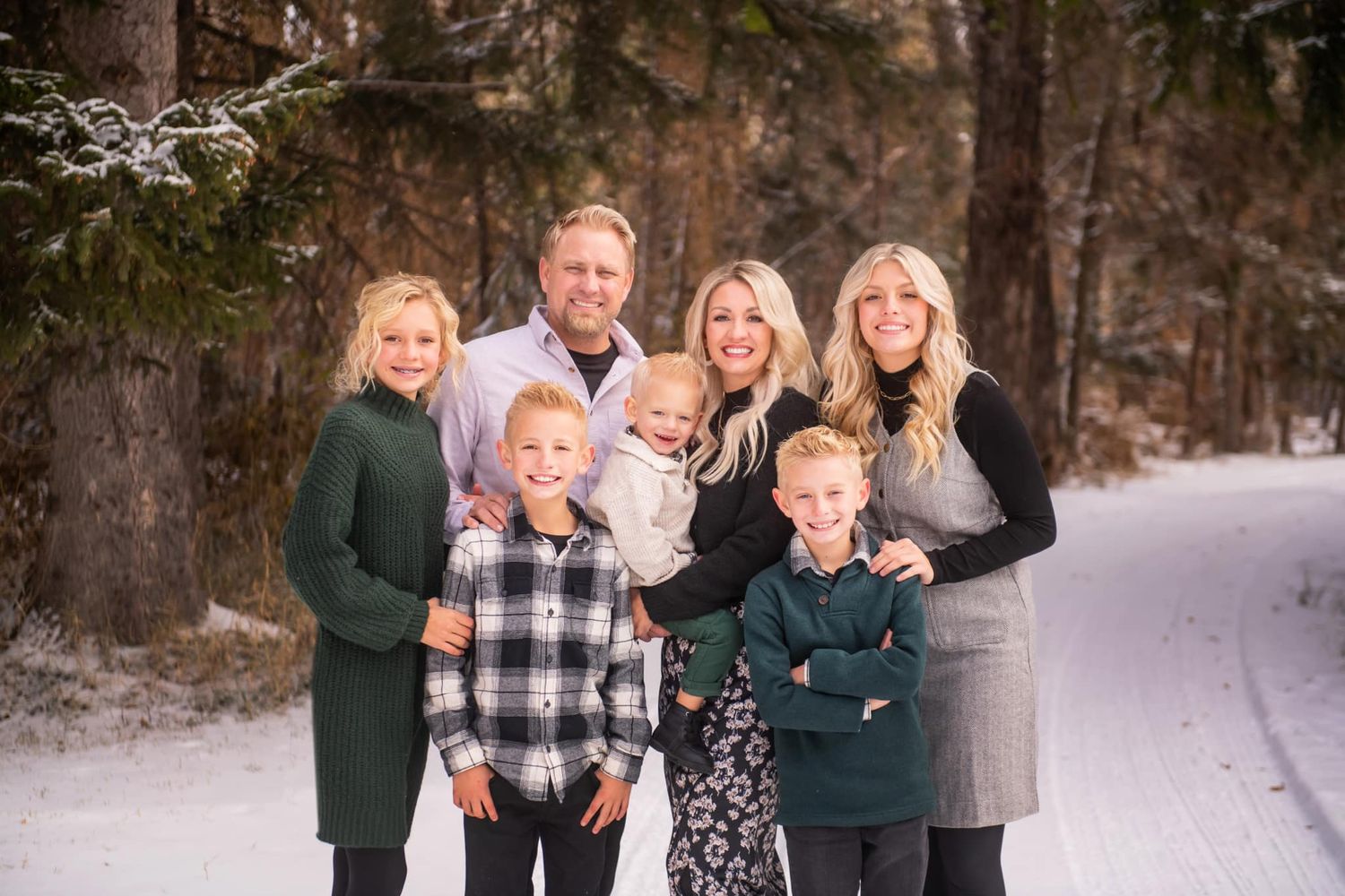 A family stands together on a snowy path in the woods wearing coordinated winter outfits in green and neutral tones.