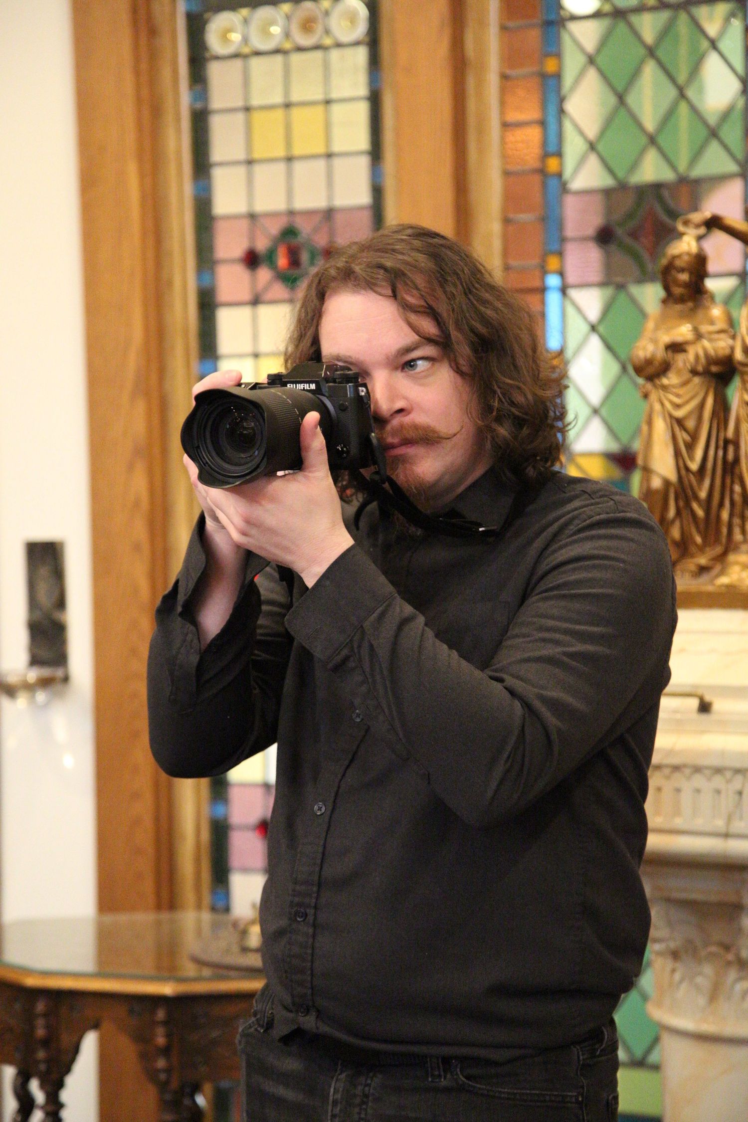 A photographer wearing dark clothing holds a professional camera while taking photos in a church interior with stained glass.