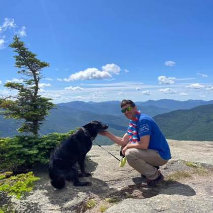 Person and black dog enjoying mountain views from a rocky summit overlook.