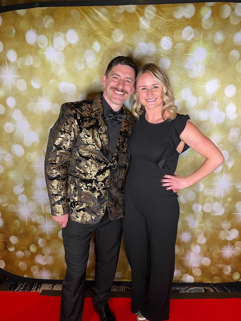 Couple posing against a sparkly gold backdrop with man wearing ornate gold patterned jacket.