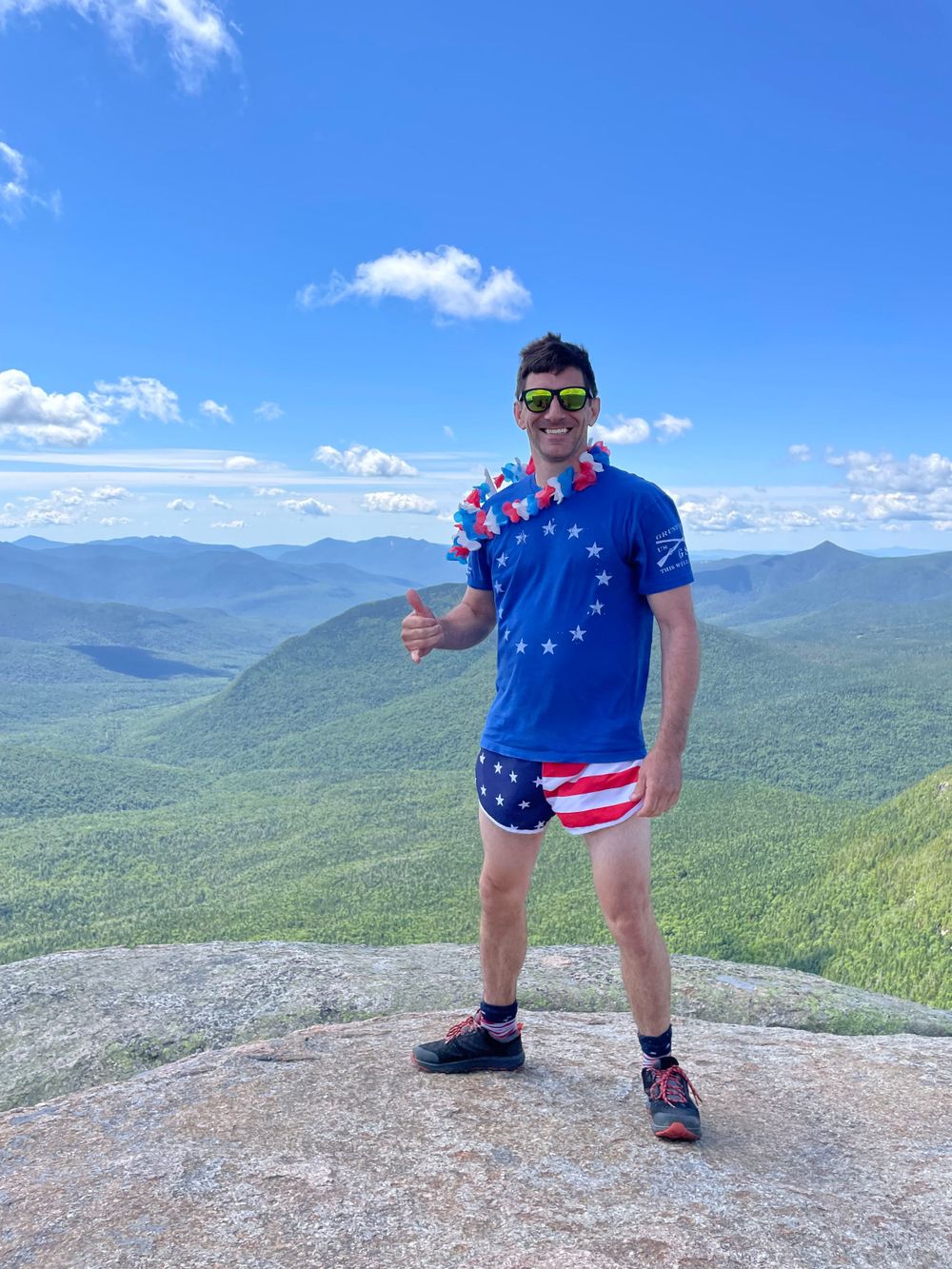 Person in patriotic American flag outfit posing on mountain summit with valley views.
