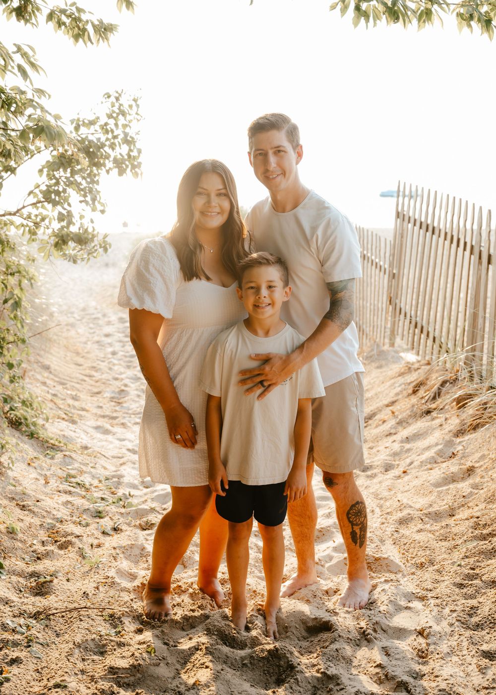 Family wearing white shirts poses together on sandy beach path with wooden fence and sunlit trees in background.