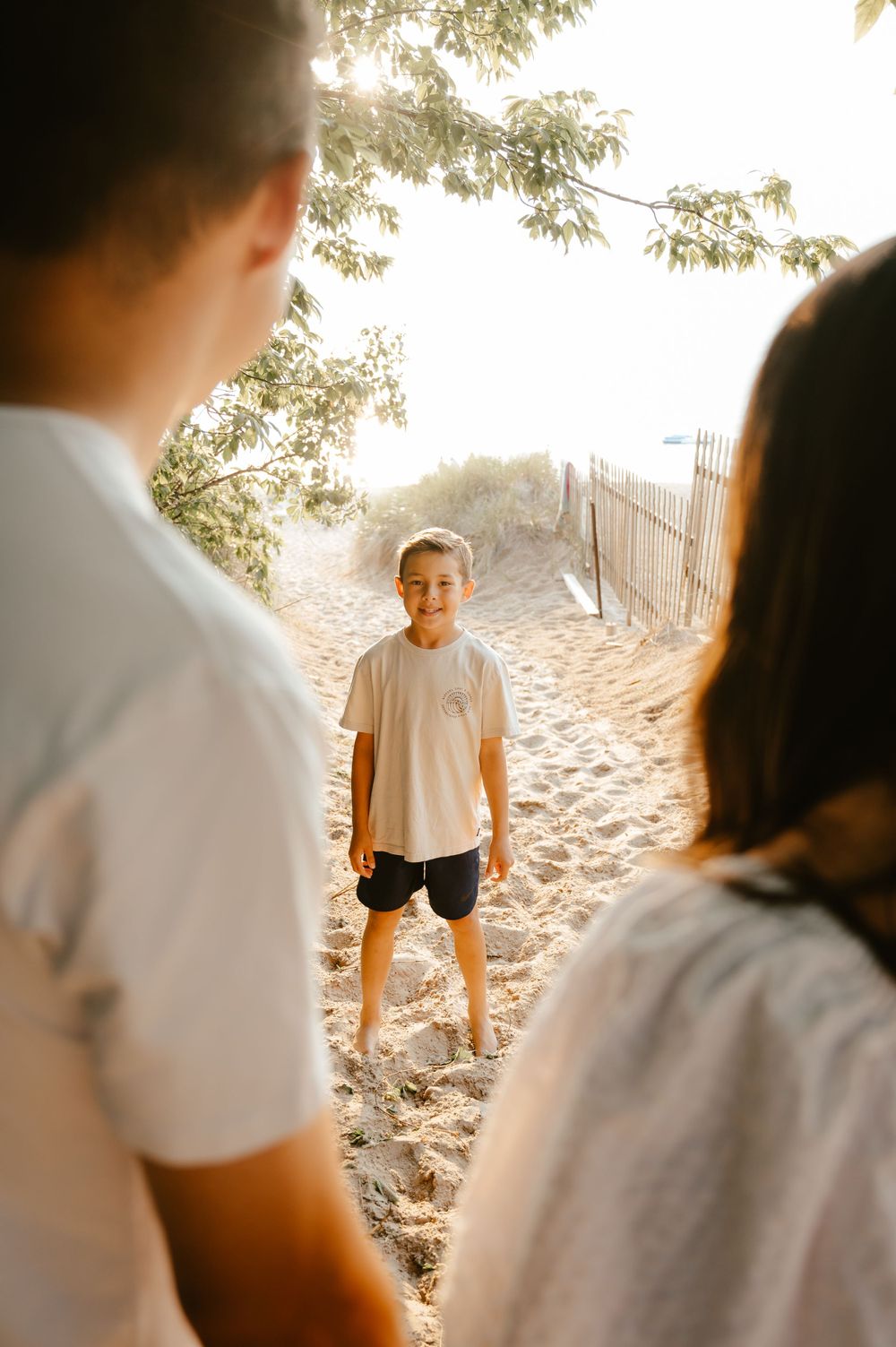 Three people in white shirts standing on a sandy beach path during golden hour with trees overhead.