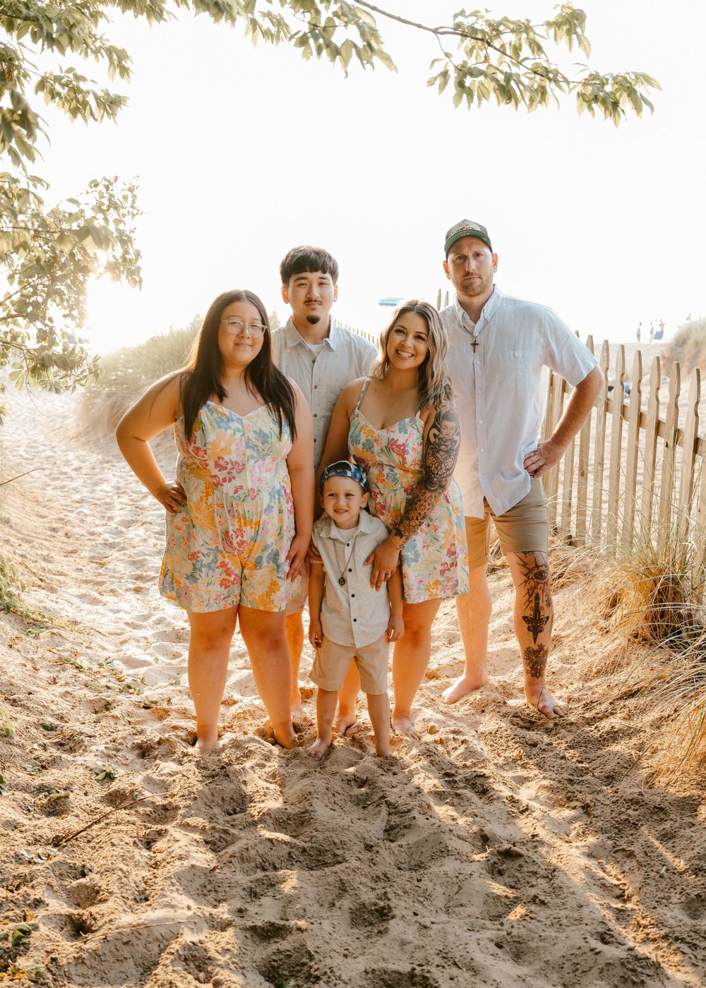 A group stands together on a sandy beach path at sunset with trees and a wooden fence in the background.