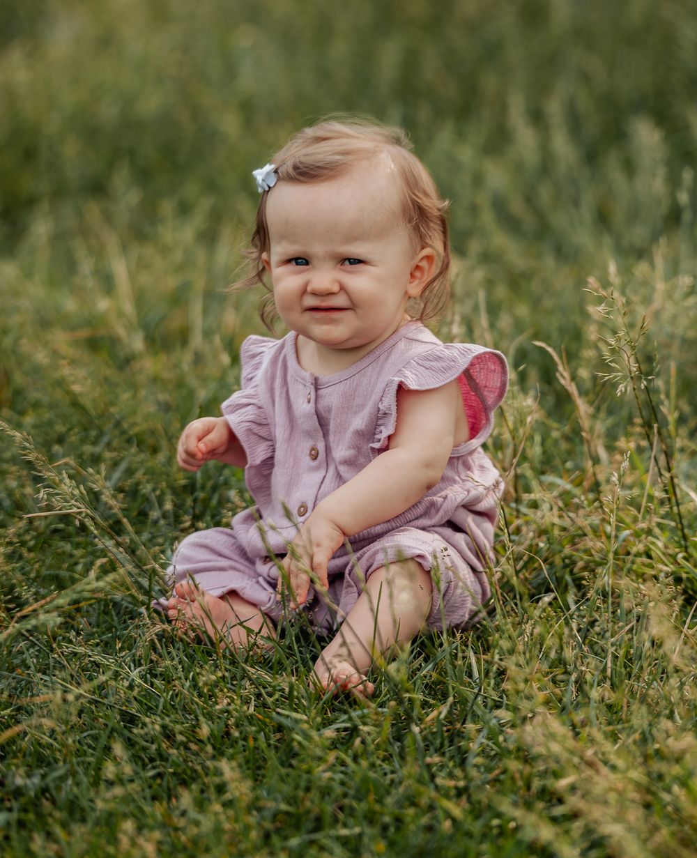 Small child in pink dress sits in tall summer grass while playing outdoors in natural lighting.