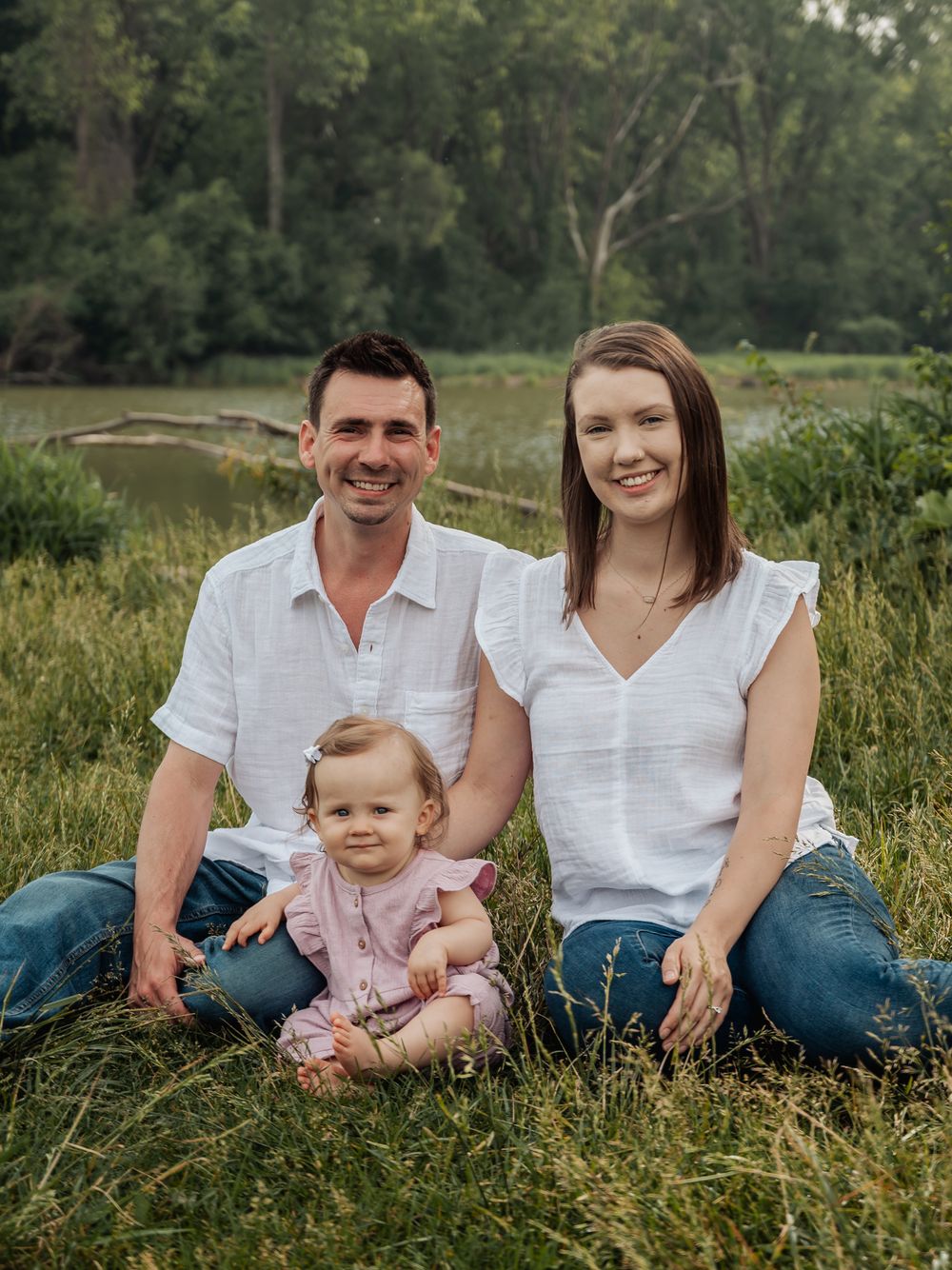 A family of three sits together in tall grass near a pond on a sunny summer day.