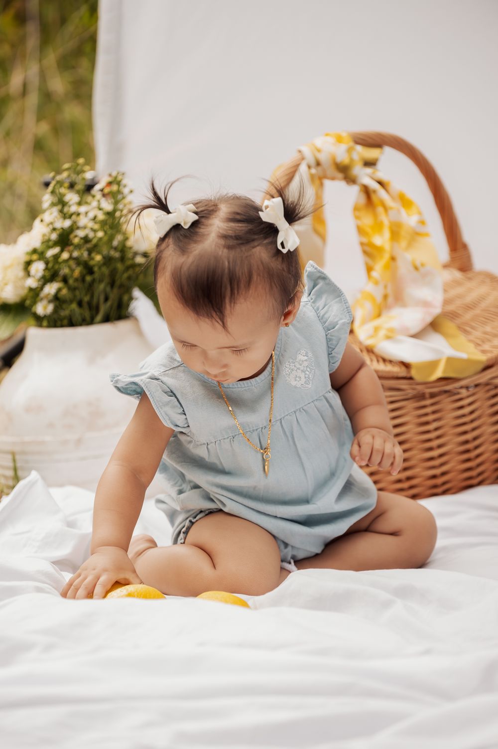Baby in blue dress sits on white blanket during outdoor picnic with wicker basket and flowers.