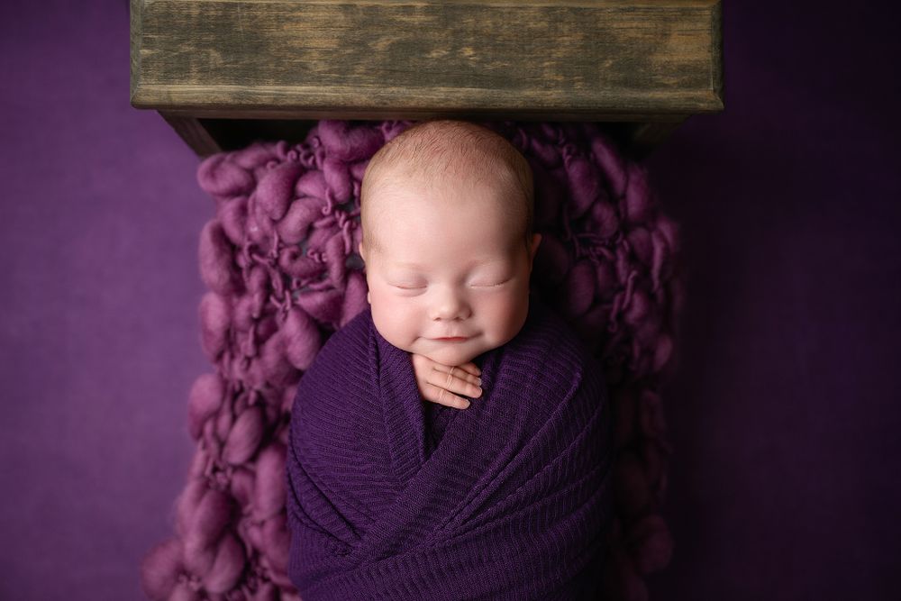 Artistic newborn portrait with purple wrap against dark wooden box and textured backdrop.