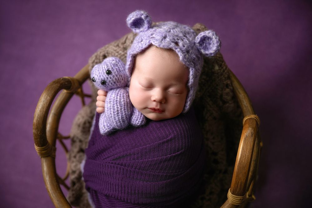Cozy newborn portrait with purple knit wrap and teddy bear hat in wooden chair.