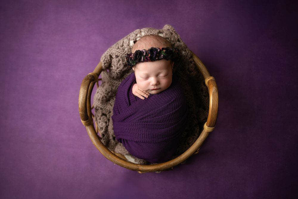 Artistic newborn photography with purple wrap in round wooden basket against dark backdrop.