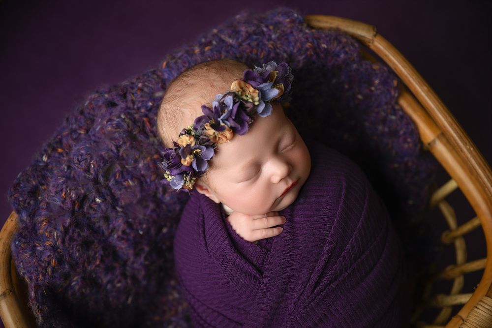 Newborn wrapped in purple fabric with floral crown resting in wooden basket against dark background.